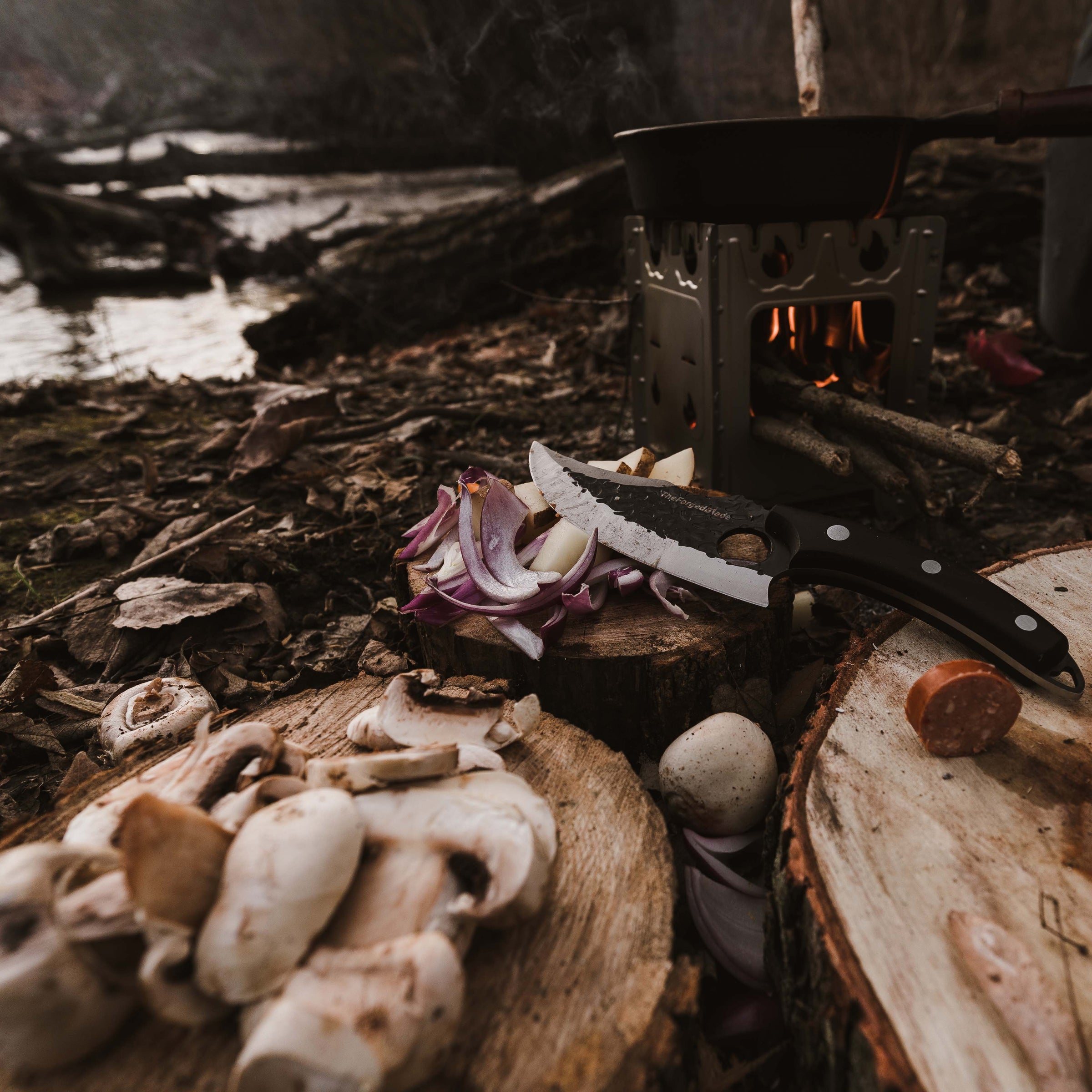 The ursa knife lying on a block of wood infront of a river and a campfire
